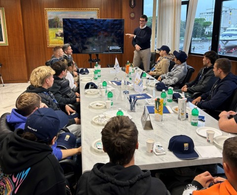 Foto interno uffici FHP: in primopiano tavolo riunioni con gli studenti seduti, sullo sfondo schermo televisivo e membro staff FHP che illustra i contenuti