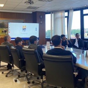 Foto interno uffici FHP: in primopiano tavolo riunioni con gli studenti seduti, sullo sfondo schermo televisivo