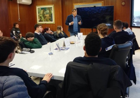 Nella foto un momento della visita: in pimo piano tavolo con attorno gli studenti, in secondo piano il relatore di fornte a una tv schermo grande